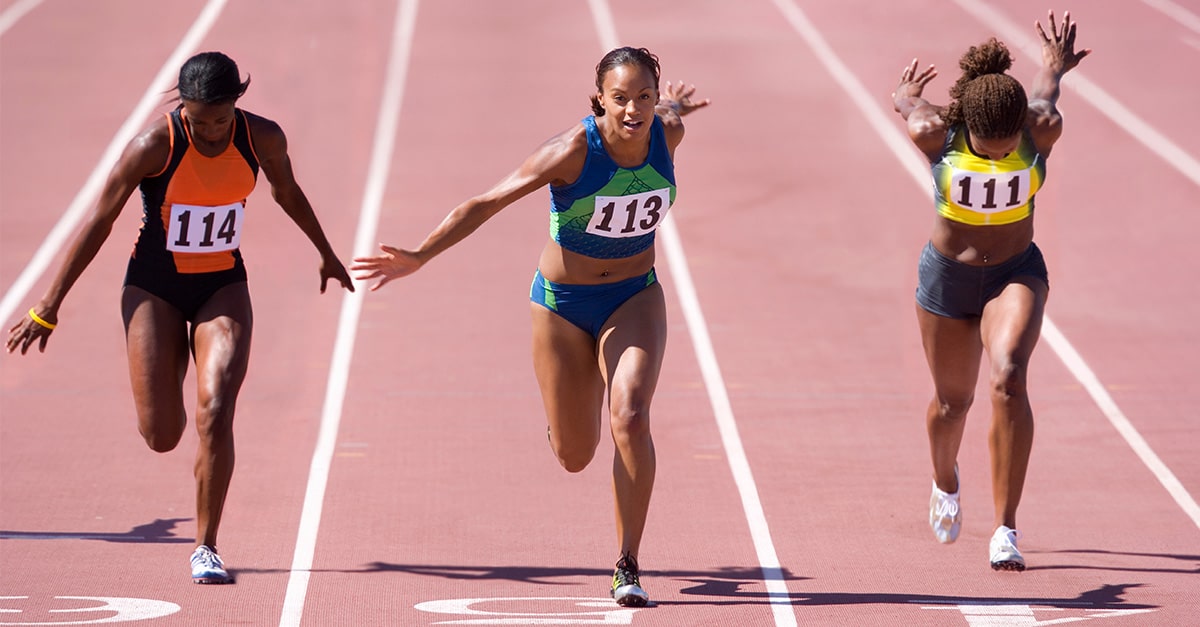 Female sprinters crossing the finish line at the end of a sprint race on a bright, sunny day at the track.
