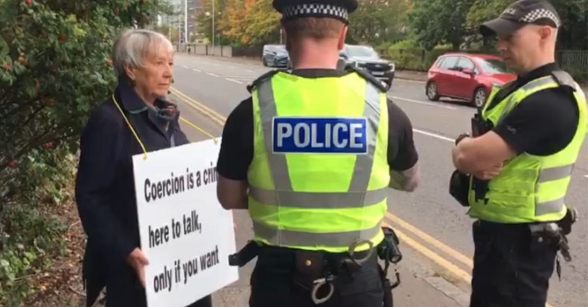 Rose Docherty, 75, talks to police who are arresting her for holding sign that says: "Coercion is a crime. Here to talk, only if you want."