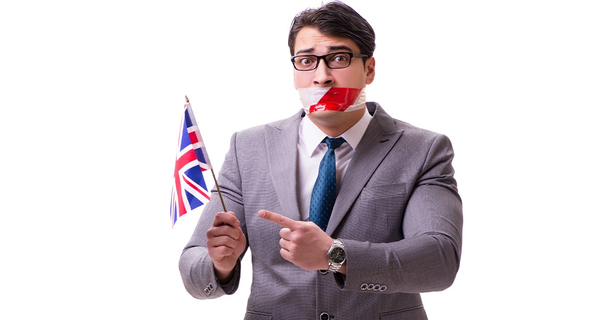 A British Man with mouth taped holds a small Union Jack flag and points to it.