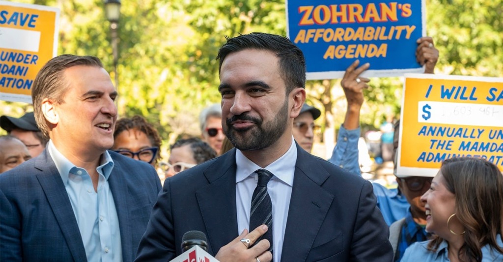 Zohran Mamdani stands in front of supporters holding signs touting his socialist affordability and savings calculator. while talking to reporters.